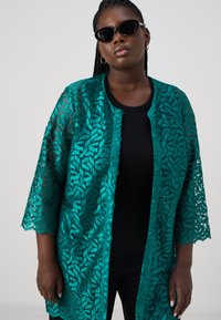 Teal kant kimono met ingewikkeld bladrandpatroon, open voorkant, 3/4 mouwen en geschulpte randen, gecombineerd met een zwarte top en zonnebril.