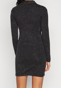 Vestido negro de manga larga con una tela texturizada que presenta un ligero brillo, llegando hasta la mitad del muslo, con cuello y silueta ajustada.