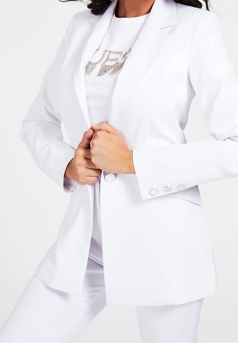 Valkoinen bleiseri, jossa on pitkät hihat, muotoiltu istuvuus, kauluskäänteet ja koristeelliset napit. Päällä valkoinen toppi, jossa on strassikuvio.