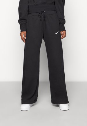 Personne portant un pantalon de jogging noir à jambes larges avec un logo Nike blanc et des baskets blanches sur un fond uni.