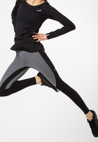 Top atletico nero con logo, abbinato a leggings color block grigi e neri. Modella in movimento, indossa scarpe sportive nere.