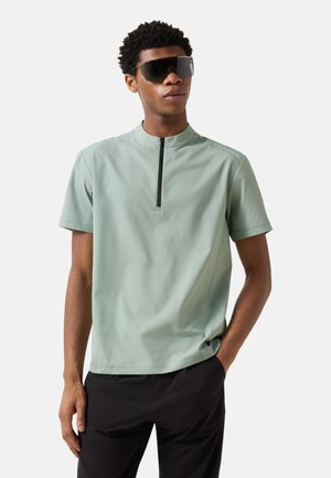 Mann trägt übergroße schwarze Sonnenbrille, hellgrünes kurzärmeliges Reißverschlusshemd und schwarze Hose, steht mit einer Hand in der Tasche.