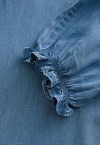Denimu audums vidēji zilā krāsā ar sakārtotu elastīgu aproču detaļām. Gluda tekstūra ar maziem tonālajiem izcēlumiem visā virsmā.