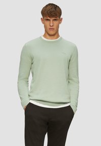 Pull en tricot vert clair avec un col ras du cou et des poignets côtelés, porté sur un sous-vêtement blanc. Le mannequin pose les mains dans les poches.