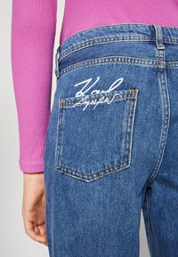 Blå denimjeans med en figursydd silhuett. Funktioner inkluderar en bakficka med vit broderad text och kontrasterande sömsdetaljer.