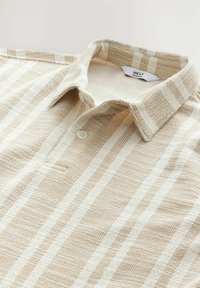 Polo majica u bež i krem prugama s dugmadi na ovratniku, teksturiranom tkaninom te malim detaljem džepa na prednjem dijelu.