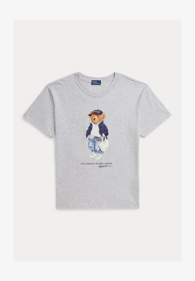 Světle šedé tričko s medvídkem Polo, který má na sobě tmavě modrou bundu, modré džíny, tenisky a čepici, drží bílou tašku, pod ním text Polo Bear od Ralph Lauren.