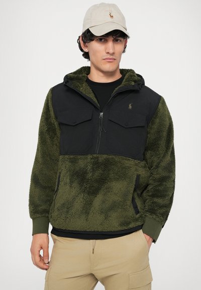 Pullover de fleece verde oliva com parte superior em nylon preto, dois bolsos no peito e capuz. Boné bege com logótipo completa o visual.