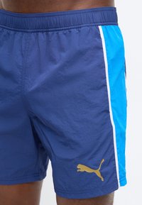 Άνδρας που φοράει μπλε σορτς Puma με γαλάζινα πλαϊνά πάνελ και λευκό περίγραμμα σε απλό φόντο.