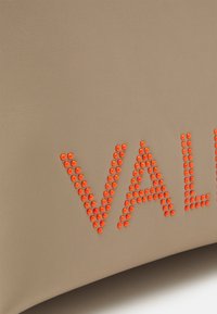 Beige läderväska med upphöjda orange nitar som formar ordet "VAL." Slät textur, rundad form, minimalistisk design och djärva accenter.