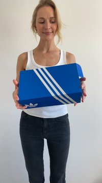 Caixa de sapatilhas azul da Adidas com três riscas brancas, apresentando uma superfície texturizada e um logo na parte da frente. Caixa segurada por uma pessoa, ligeiramente inclinada.
