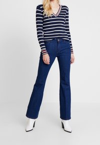 Marineblaue und weiße, gestreifte Langarmshirt mit V-Ausschnitt und gerippter Struktur, kombiniert mit ausgestellten, dunkelblauen Jeans und weißen Stiefeln mit Absatz.