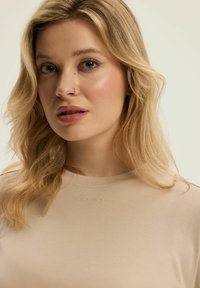 Jasno beżowa bluzka z okrągłym dekoltem o subtelnej teksturowanej powierzchni i haftowanym logo na klatce piersiowej, noszona przez osobę z falującymi, blond włosami.