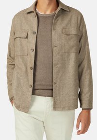 Brun herringbone skjortjacka med krage, två bröstfickor och knäppningar. Bärs över en brun tröja och ljusa byxor.