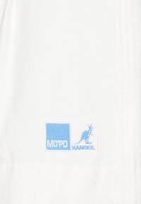 Biała tkanina z małą, niebieską prostokątną metką z napisem "MO'PD" i logo kangura, wskazującą markę Kangol.