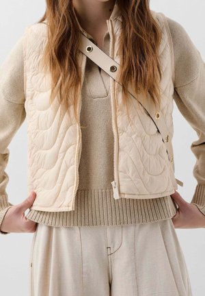 Femme portant un pull en maille beige, un gilet matelassé crème, un pantalon clair plissé, avec une bandoulière crème traversant la poitrine.