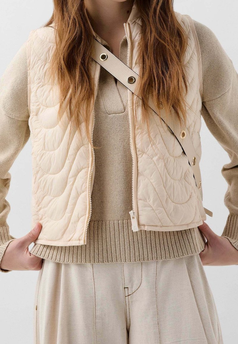 Femme portant un pull en maille beige, un gilet matelassé crème, un pantalon clair plissé, avec une bandoulière crème traversant la poitrine.