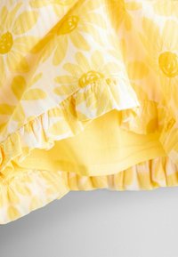 Material floral galben și alb cu margini franjurate, prezentând o prim-plan detaliat al tivului și cusăturilor stratificate.