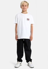 Hvit bomull t-skjorte med et lite trykt grafikk på venstre bryst, paret med svarte cargo-bukser og hvite joggesko med grønne detaljer.