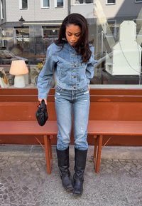 Veste en denim bleu clair avec un design court, portée sur un jean bleu clair. Présente des bottes noires jusqu'aux genoux et tient un sac noir.