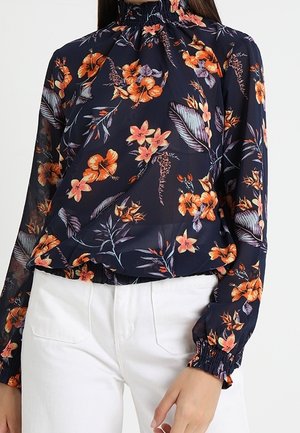 Granatowa bluzka z wysokim marszczonym kołnierzem i mankietami, z pomarańczowo-różowym kwiatowym nadrukiem, noszona z białymi spodniami.