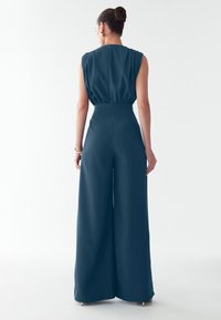 Tume teal ühes tükis, millel on volditud õlgade disain, kitsendatud vöökoht ja laiad püksid. Sile tekstuur, seljaosa lukuga.