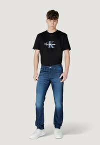 Camiseta negra con el logo de Calvin Klein Jeans, combinada con jeans de lavado medio. El modelo lleva zapatillas blancas. El fondo es de un gris claro.