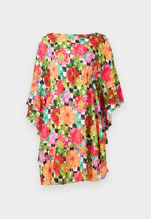 Žena stojí v voľných kvetovaných šatách po kolená s oranžovými, ružovými a zelenými kvetmi a čiernych sandáloch na podpätku.