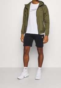 Giacca leggera verde oliva, T-shirt bianca con righe nere, pantaloni corti neri e sneakers bianche. Caratteristiche: chiusura con zip e cappuccio regolabile.