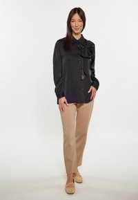 Blusa nera in satin con dettaglio a fiocco, davanti con bottoni, maniche lunghe, abbinata a pantaloni beige su misura e mocassini marroni chiari.