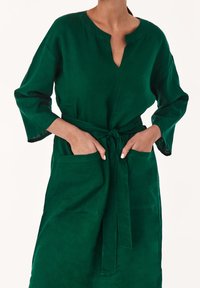 Zelené lněné šaty s výstřihem do V, dlouhými rukávy a nudlí v pase. Obsahují dvě přední kapsy a hladký, strukturovaný povrch.