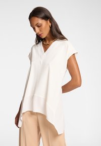 Hvid, letvægts bluse med V-hals og korte ærmer; asymmetrisk kant giver fluiditet; bæres med beige bukser.