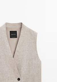 Beige linnen mouwloos vest met diepe V-hals en enkele zwarte knoopsluiting, met een Massimo Dutti-label aan de binnenkant van de kraag.