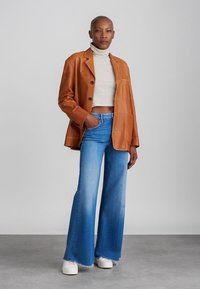 Hnědý kožený oversized blazer, bílý žebrovaný rolák, světle modré rozšířené džíny a bílé boty na neutrálním pozadí.