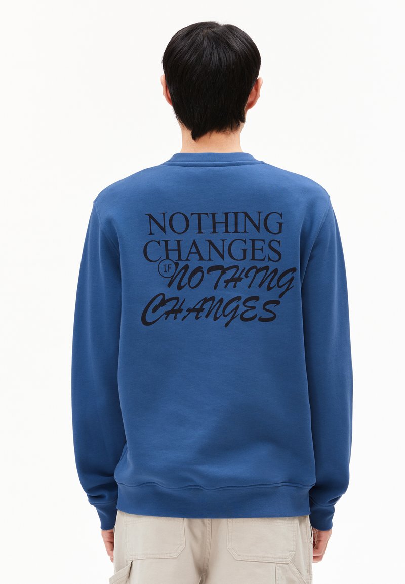 Blauwe sweatshirt met zwarte tekst op de achterkant die leest: "ALS ER NIETS VERANDERT, VERANDERT ER NIETS." Zachte stof, ronde hals, lange mouwen.