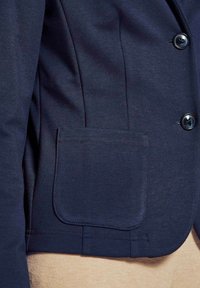 Marineblauer Blazer mit glatter Textur, funktionalen Knöpfen, einer aufgesetzten Tasche und taillierten Nähten an den Seiten.