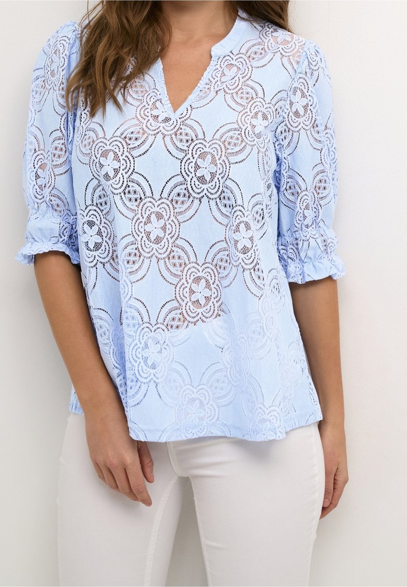Haut en dentelle bleu clair avec motif floral, manches courtes bouffantes et encolure en V ; présente une texture transparente et des détails découpés complexes.