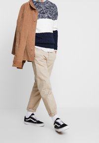 Brun manchesterjacka, marinblå och vit stickad tröja, beige byxor, svart- och vitrandiga sneakers, vita strumpor, avslappnad passform, prydlig design.