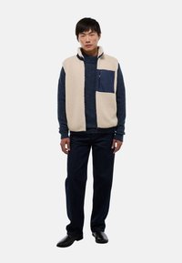 Gilet en polaire beige clair avec un col haut et une poche contrastante marine, porté sur un pull en laine texturé bleu foncé et un pantalon en jean foncé.