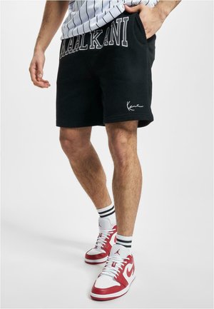 Black cotton shorts with "Kaal Kani" printed on the waistband, featuring a slim fit and embroidered logo detail. Paired with red and white sneakers.