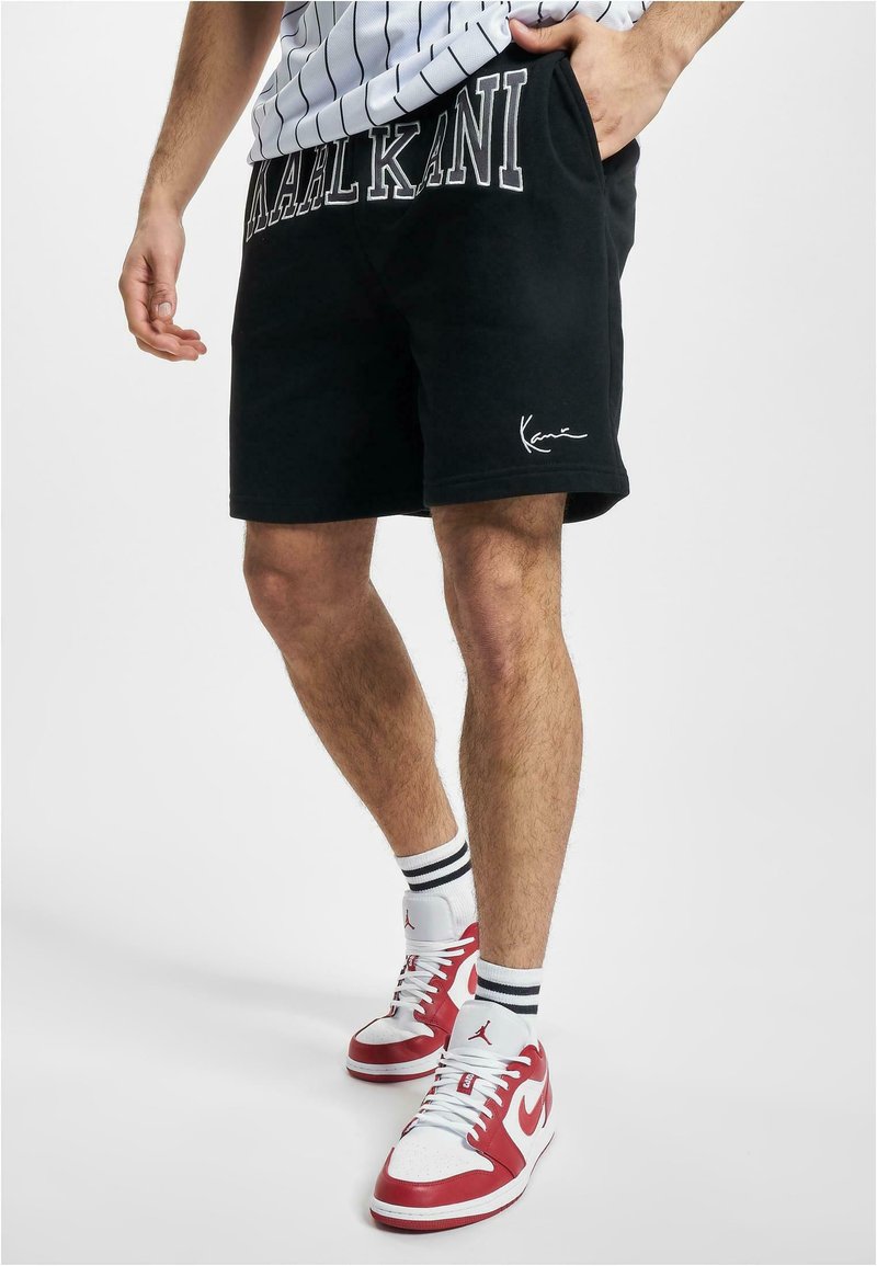 Crne pamučne kratke hlače s natpisom "Kaal Kani" na pojasu, sa slim fit krojem i vezenim logotipom. U paru s crvenim i bijelim tenisicama.