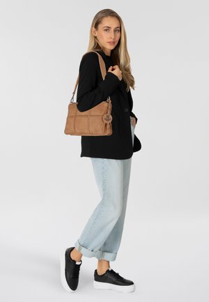 Borsa in pelle marrone con design trapuntato, dotata di un charm a forma di ancora in argento. Modello che indossa un blazer nero, jeans chiari e sneaker nere.