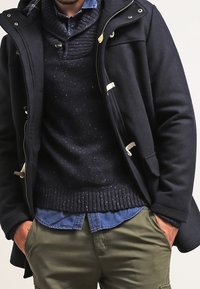 Abrigo de lana azul marino con cierres de toggles, sobre un suéter de punto oscuro, en capas sobre una camisa de botones azul, combinado con pantalones oliva.