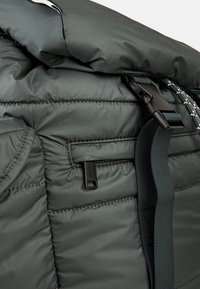 Dunkelgrüner gepolsterter Rucksack mit einem Rolltop-Verschluss, verstellbarem Tragegurt und einer Reißverschluss-Tasche mit einem rechteckigen Ziehband.