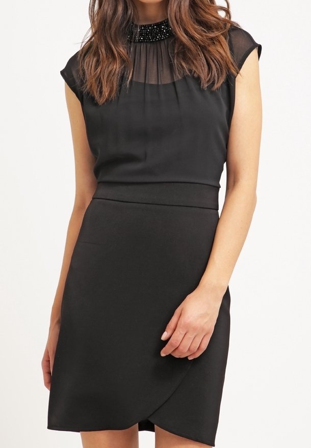 Femme portant une robe noire sans manches avec un décolleté transparent orné de perles et une taille cintrée, debout devant un fond clair uni.