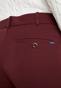Burgundské kalhoty vyrobené z hladkého materiálu, vybavené zadní kapsou se zapínáním na knoflík a malou modrou etiketou s nápisem "Polo."
