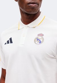 Homme portant un polo blanc avec un liseré jaune, logo du Real Madrid sur le côté gauche de la poitrine, et logo Adidas sur le côté droit.