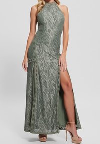 Olivengrøn maxikjole med halterneck, geometrisk udskæringsmønster, sideslids og blondeaccenter. Båret med beige højhælede sandaler.