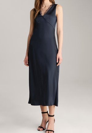 Kobieta w granatowej, satynowej sukience midi bez rękawów z koronką przy dekolcie w kształcie litery V oraz w czarnych sandałach na wysokim obcasie z paskiem wokół kostki.