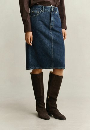 Person trägt einen knielangen dunkelblauen Jeansrock, ein dunkelbraunes langärmeliges Hemd und dunkelbraune Wildleder-Kniehochstiefel mit kleinem Absatz.